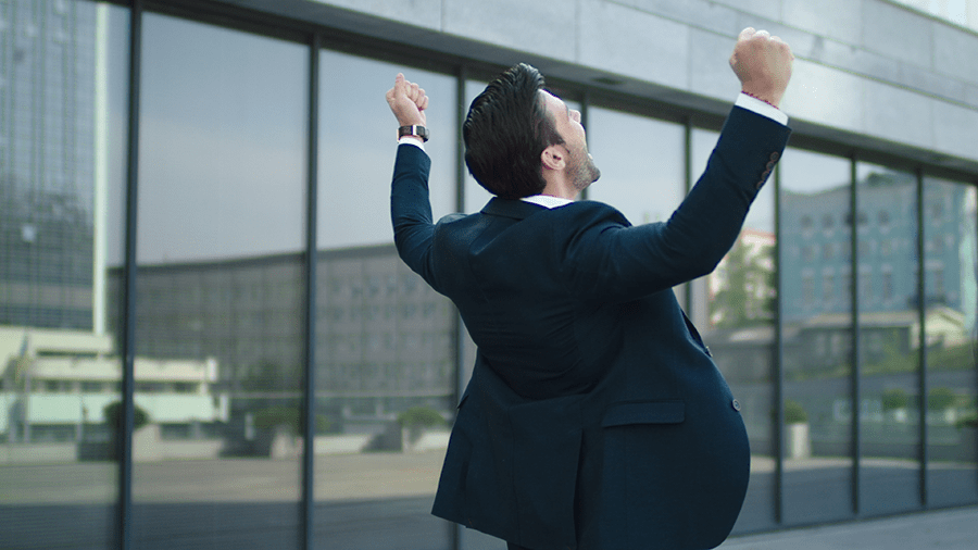 A man in a business suit is standing outside with his arms raised in a victorious or celebratory gesture.