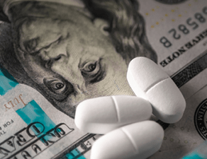 The image is of various denominations of U.S. currency, white pills, and a medical syringe, suggesting a theme related to healthcare costs or pharmaceuticals.
