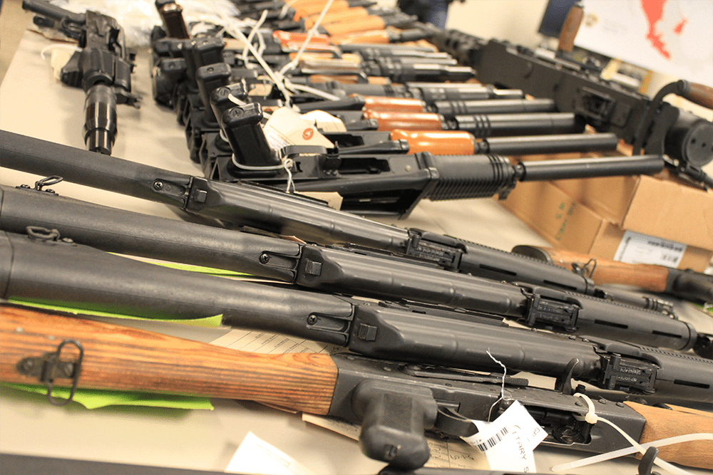 The image shows a collection of rifles and shotguns displayed on a table, possibly in a law enforcement or military context.