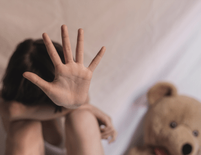 A child sitting beside a teddy bear is holding up their hand as if to signal stop or to shield their face.