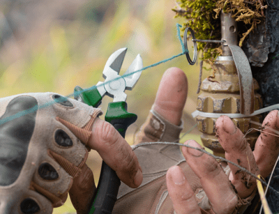 Two gloved hands use pliers to cut wire on a camo-clad booby trap in a natural outdoor setting.
