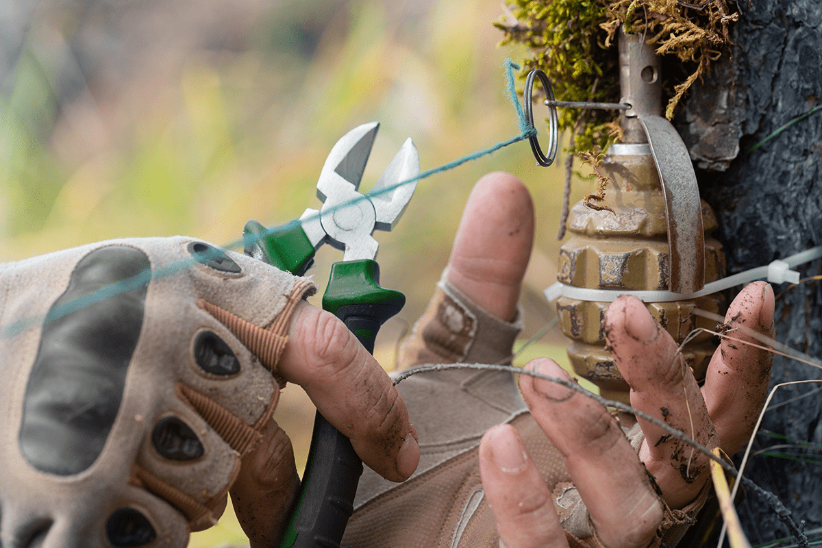 Two gloved hands use pliers to cut wire on a camo-clad booby trap in a natural outdoor setting.