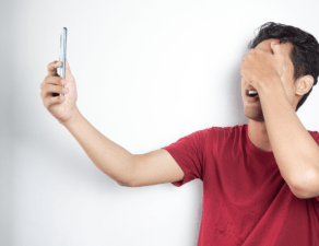 A man in a red shirt holds a phone at arm's length, seemingly frustrated or covering his face with his other hand, possibly reacting to the content on the screen.