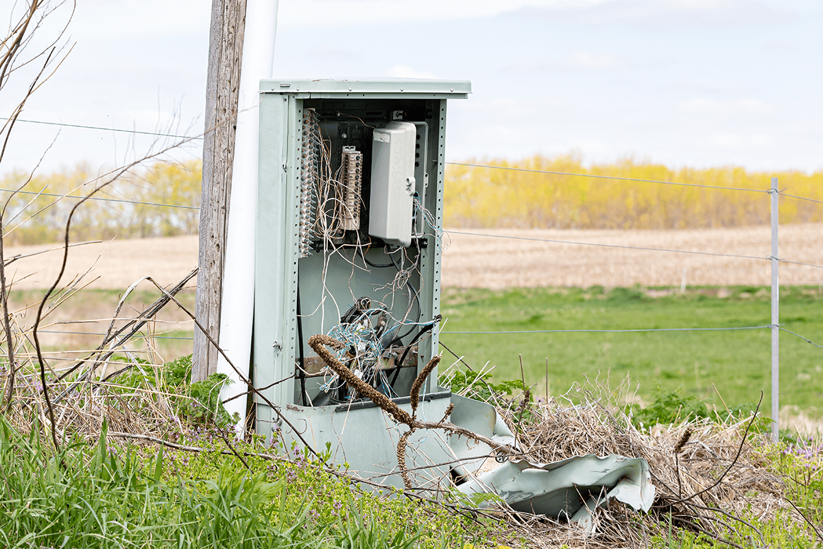 An open, damaged utility box with exposed wires stands in a grassy area near a field.