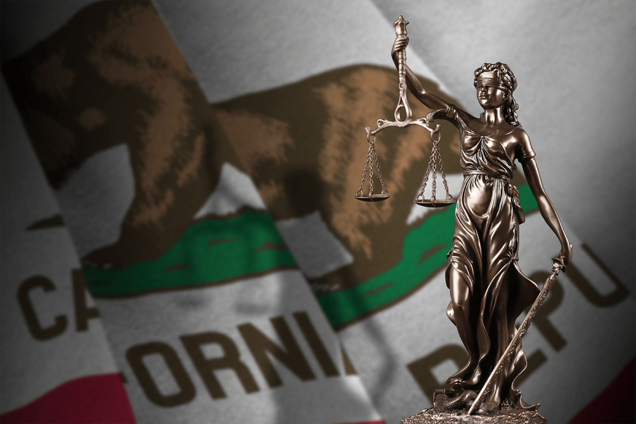 A statue of Lady Justice is in the foreground with the flag of California in the background.