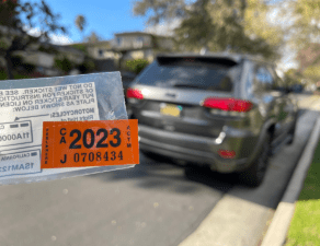 A vehicle registration sticker for 2023 is in focus in front of a blurred car parked on a suburban street.