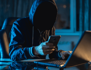 A hooded individual with obscured face is using a smartphone and laptop in a dark room, suggesting secretive or illicit activity.