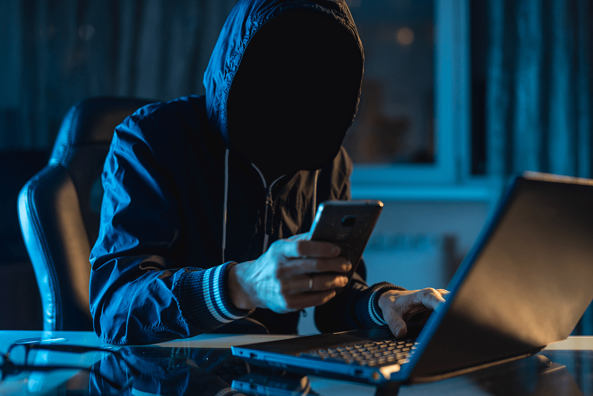 A hooded individual with obscured face is using a smartphone and laptop in a dark room, suggesting secretive or illicit activity.